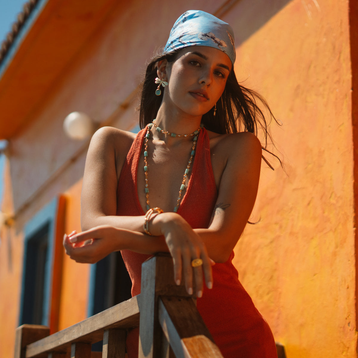 Model leaning on a wooden balcony rail in a red dress and blue headscarf, wearing the Spiritual Dragonfly aqua earrings in the sun