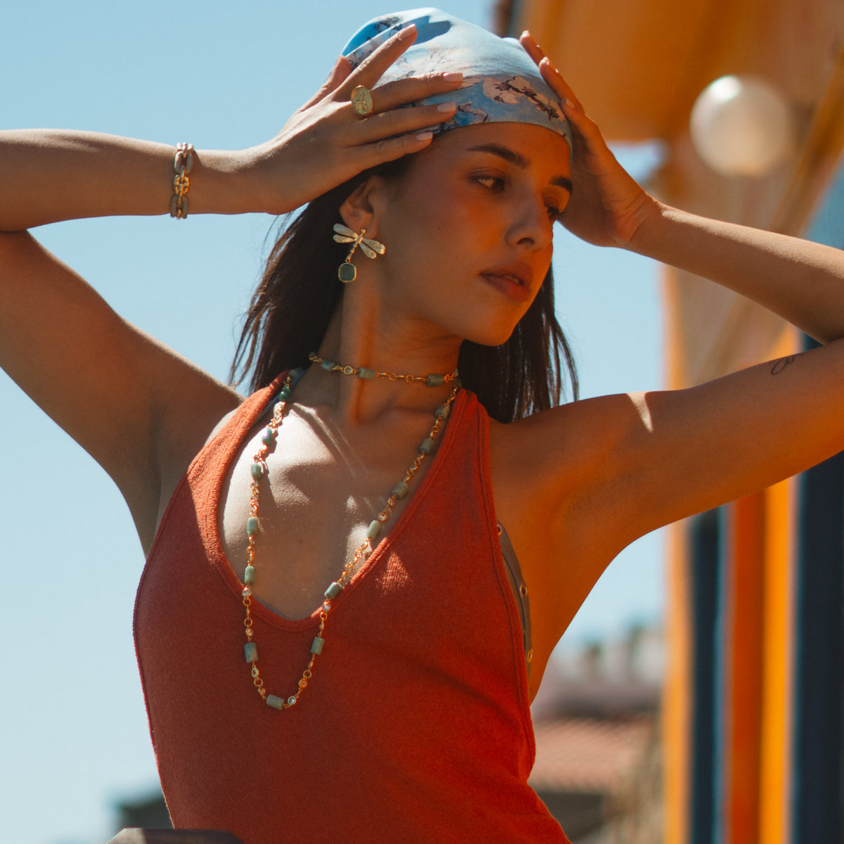 Model with both hands on her blue headscarf against a clear sky, wearing the gold dragonfly earrings and long beaded necklaces