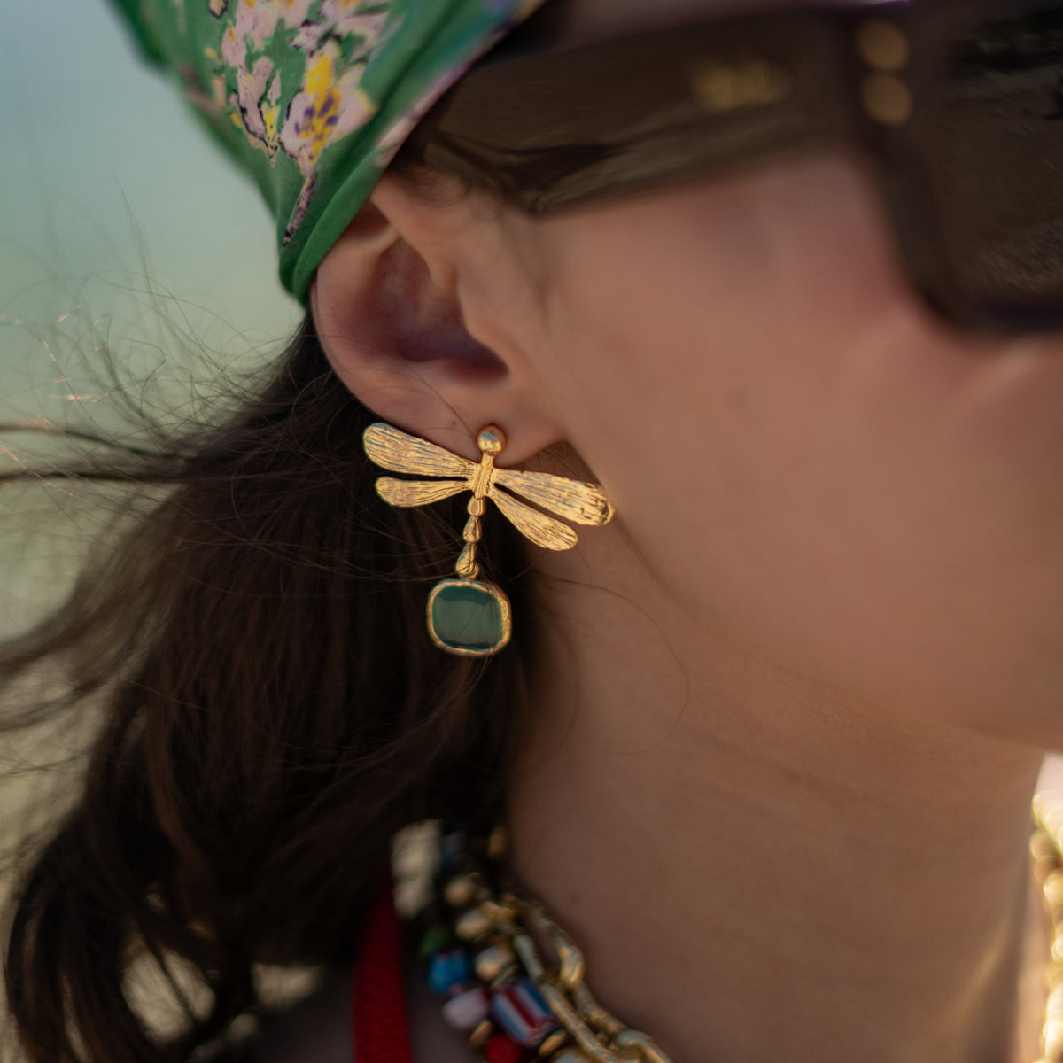 Close view of the gold dragonfly earring with its aqua cat's eye drop, worn with a floral green headscarf and colorful necklaces