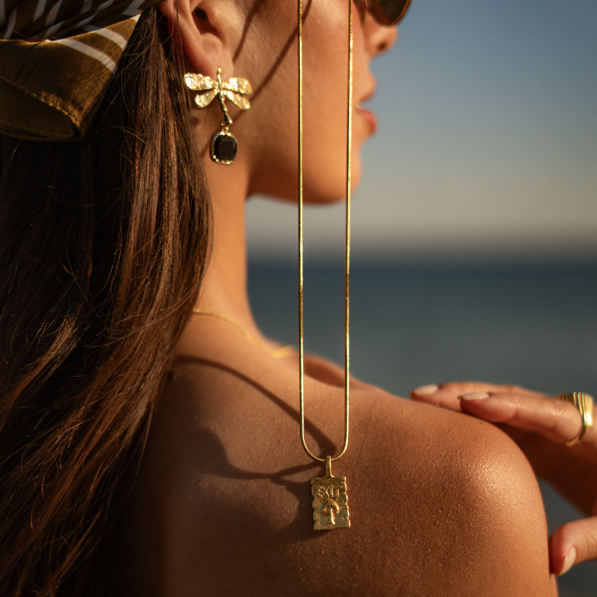 Model by the sea wearing the gold dragonfly black cat's eye earring with a long gold chain and engraved tablet pendant