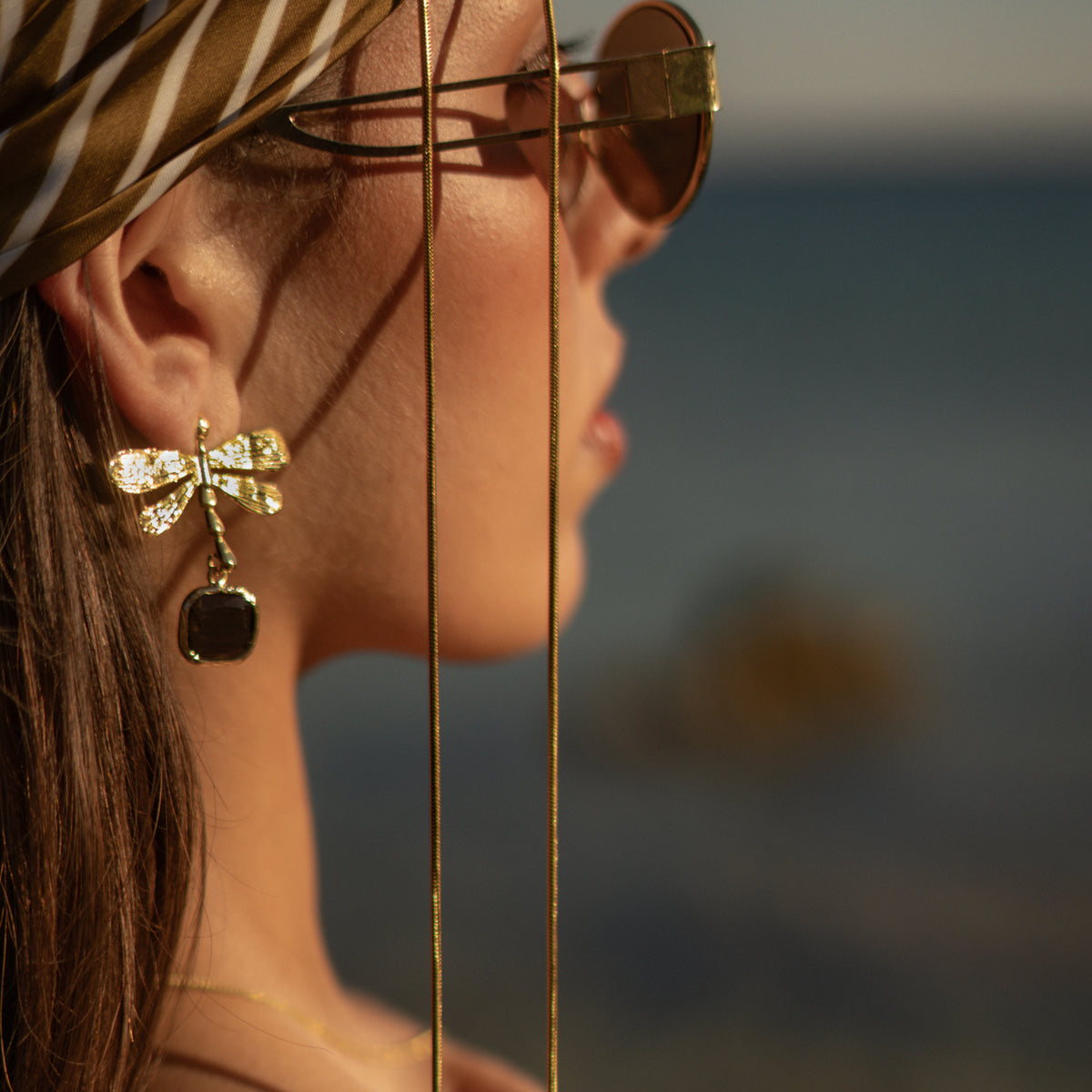 Profile of a model in sunglasses and a striped headscarf wearing the gold dragonfly earring with its black cat's eye drop