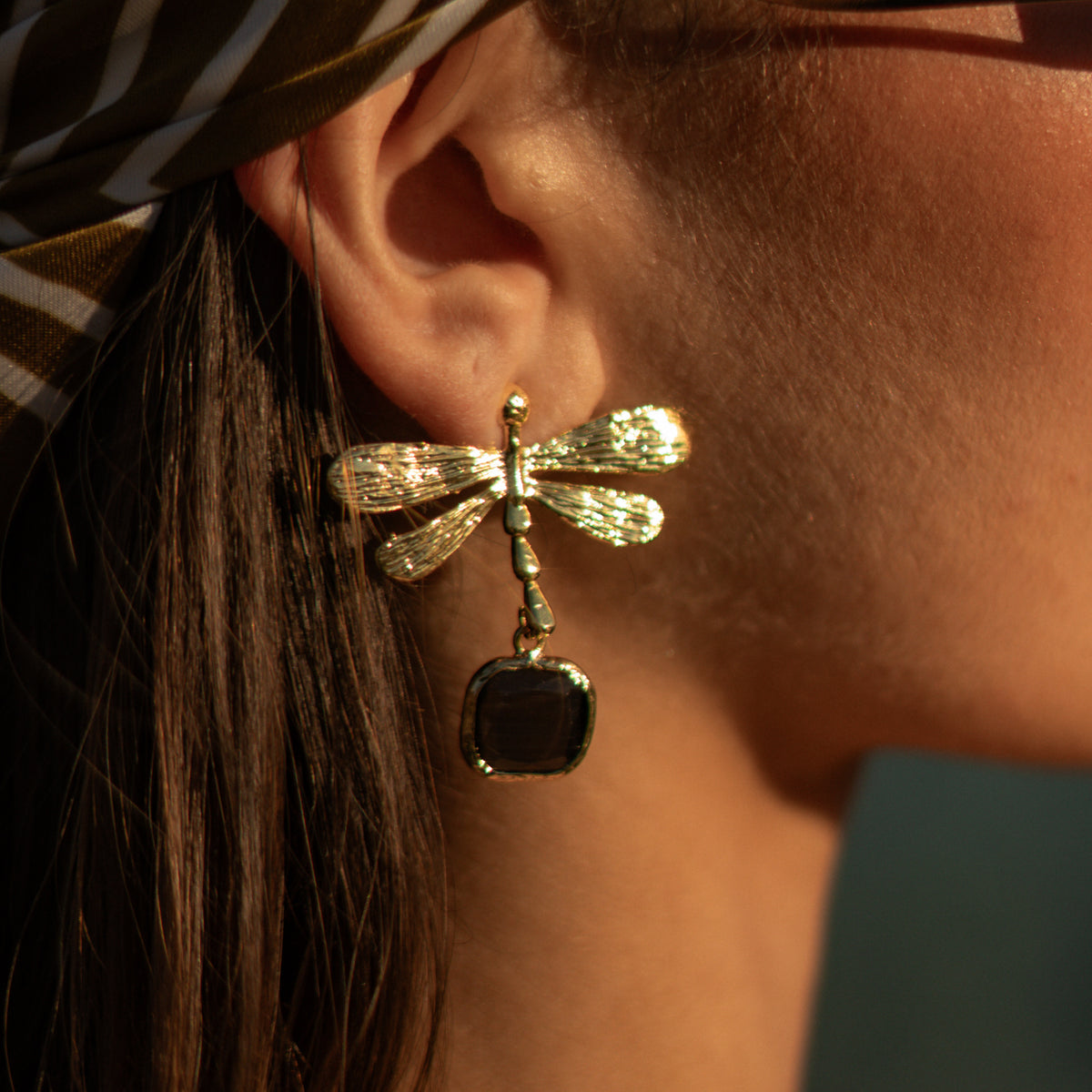 Close side view of the Spiritual Dragonfly black cat's eye earring on the ear in warm evening light, below a striped headscarf