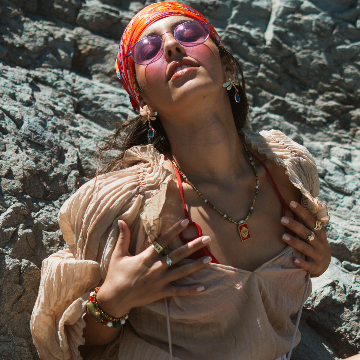 Model in an orange headscarf and sheer blouse against grey rocks, wearing the blue dragonfly earrings with a beaded pendant necklace