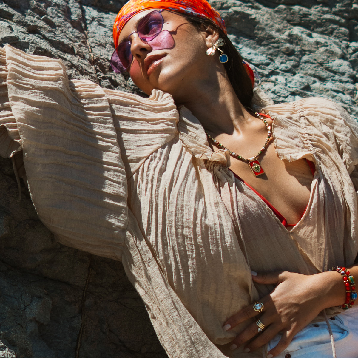 Model reclining on sunlit rocks in tinted sunglasses, wearing the Spiritual Dragonfly blue earrings and layered necklaces