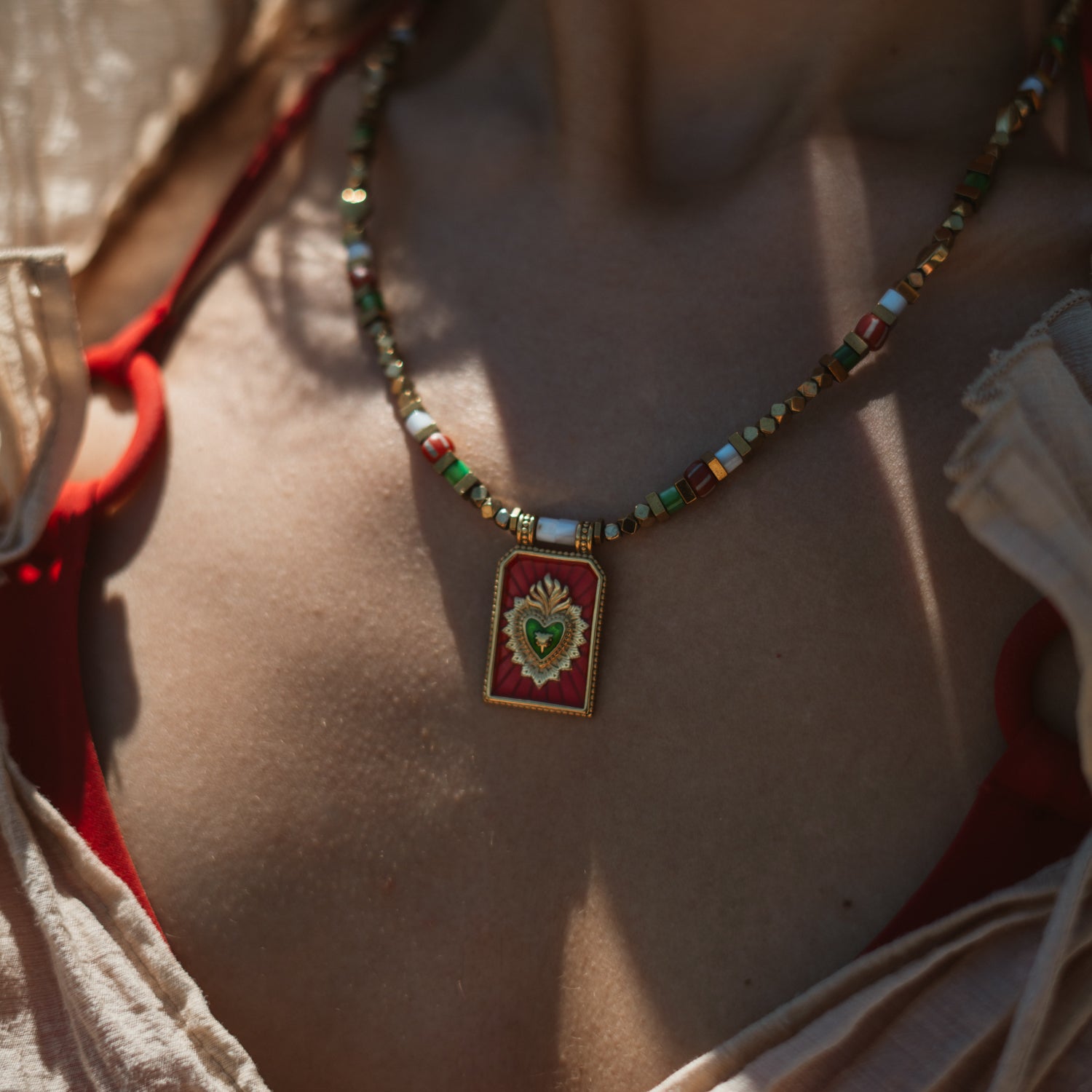 A close-up lifestyle shot of a person wearing a colorful beaded necklace with a red and green heart-shaped pendant, set against warm sunlight and neutral-toned clothing.