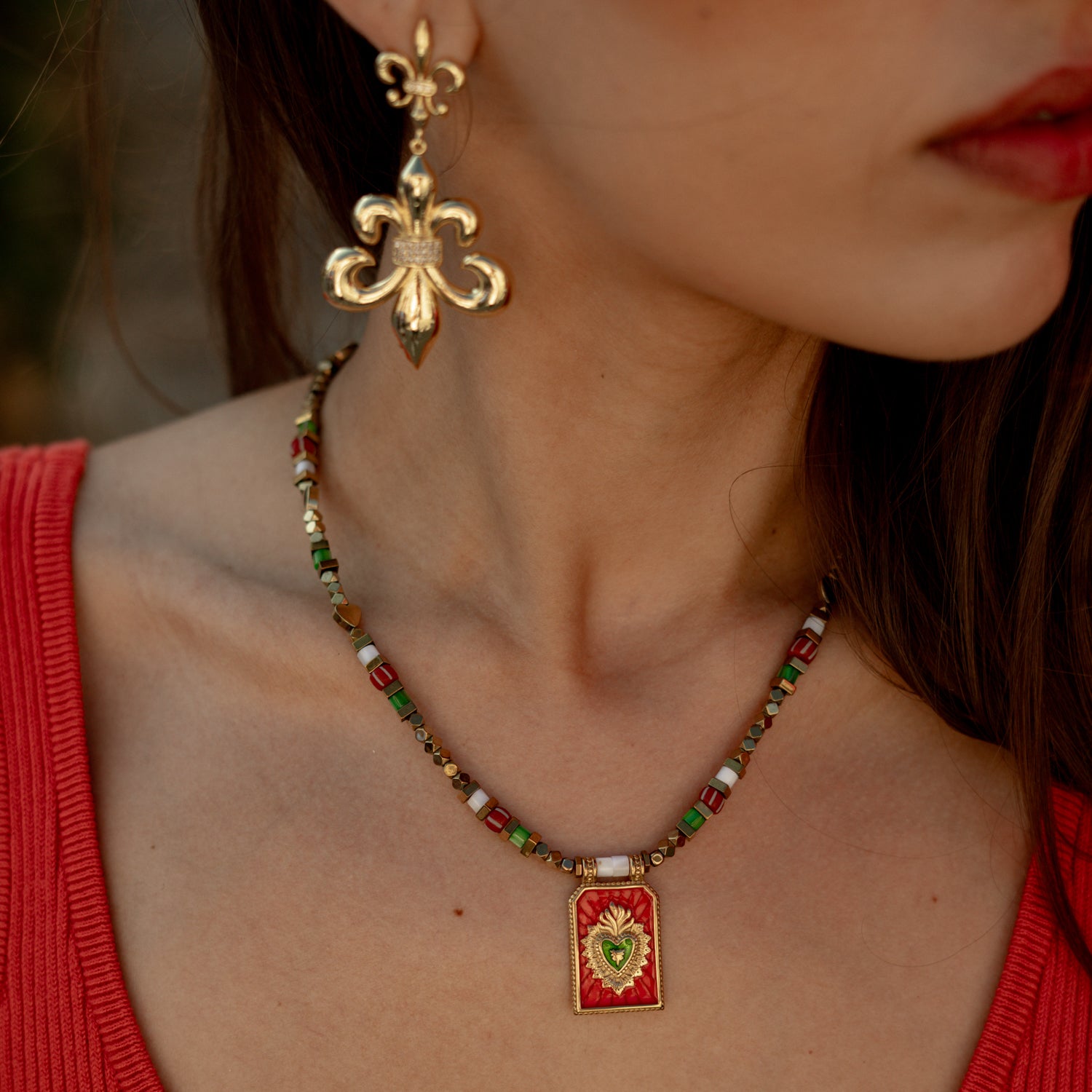A close-up lifestyle shot of a woman wearing a red and gold pendant necklace and a matching gold fleur-de-lis earring. The necklace features a red rectangular pendant with a gold Sacred Heart design, and a beaded chain with red, green, white, and gold beads.