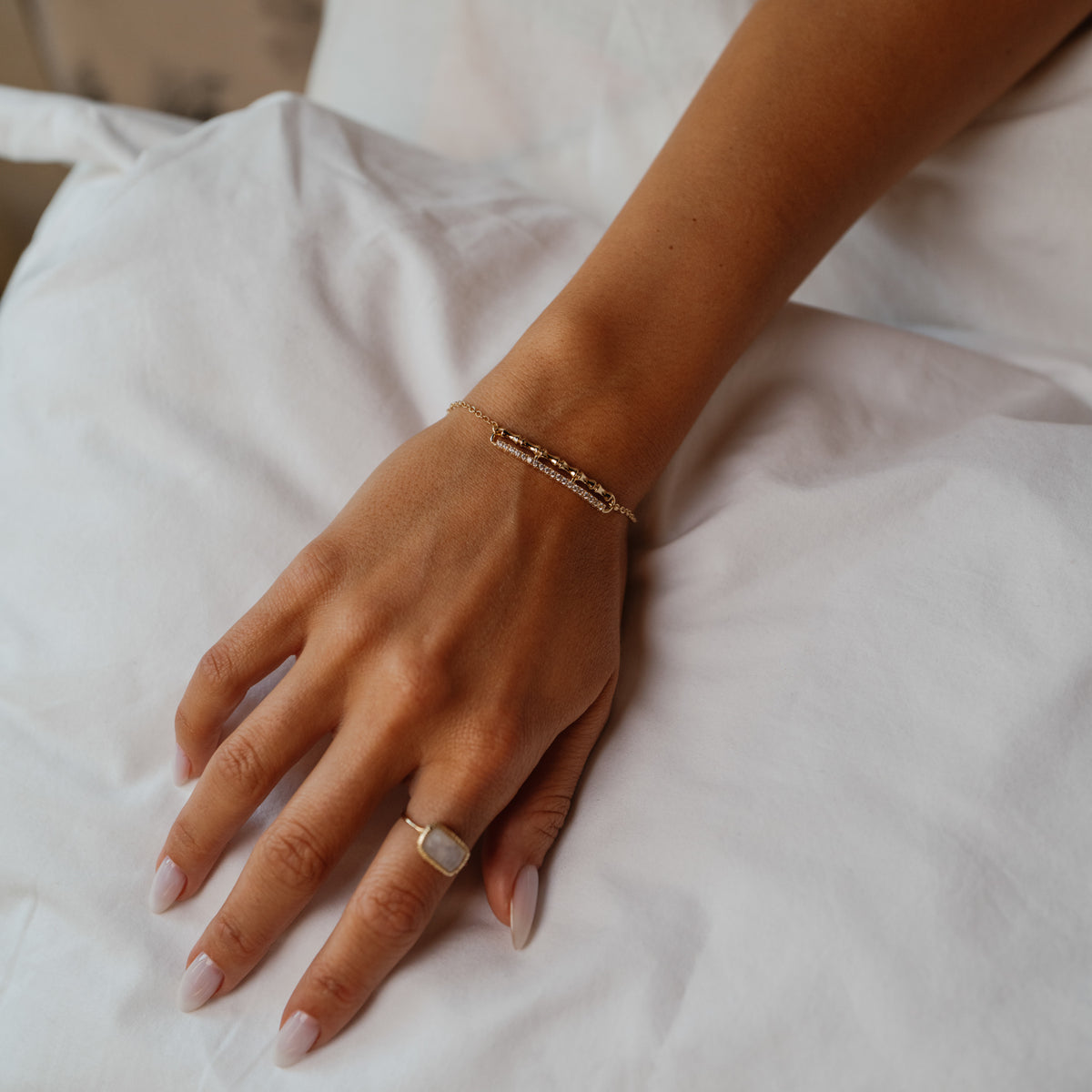 Arm across white bedding, the Golden Bamboo gold zircon bar bracelet on the wrist and a gold stone ring on the hand