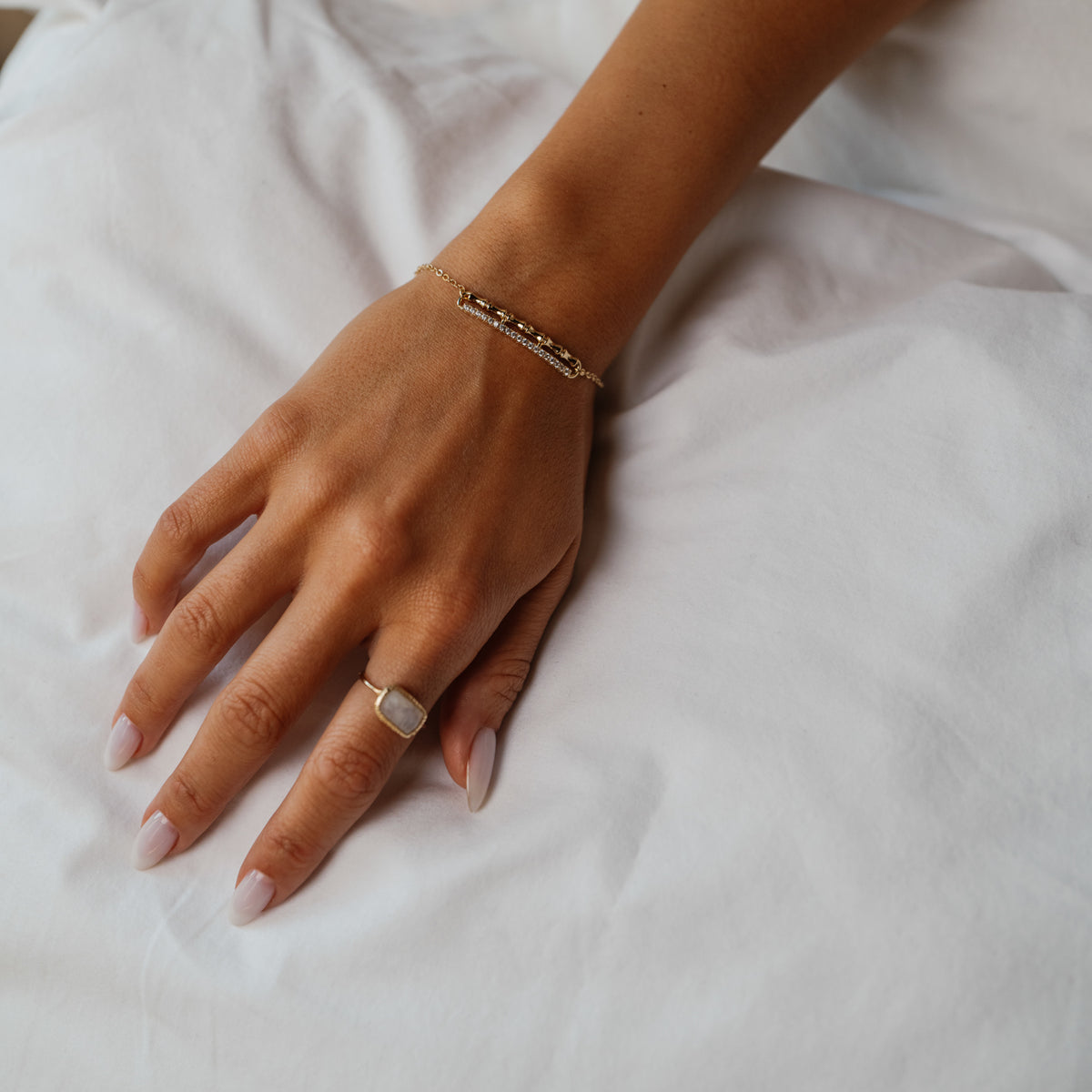 Hand resting on white linen wearing the Golden Bamboo gold bar bracelet with a gold ring set with a square milky stone