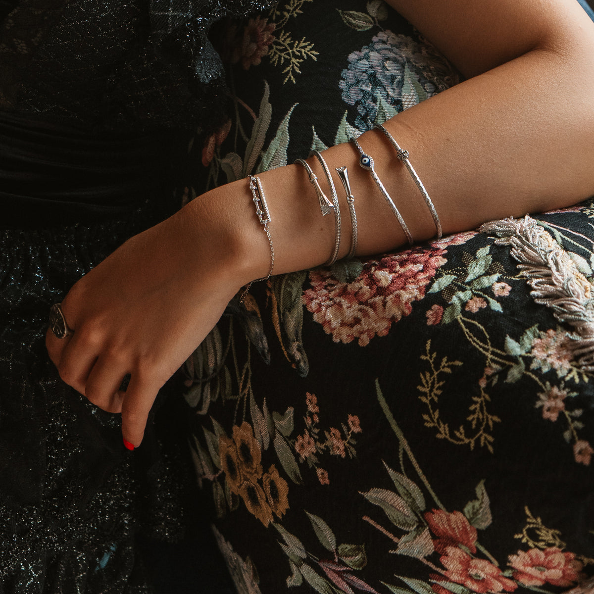 Arm resting on a floral cushion, the Three Lights silver bar bracelet stacked with slim silver bangles