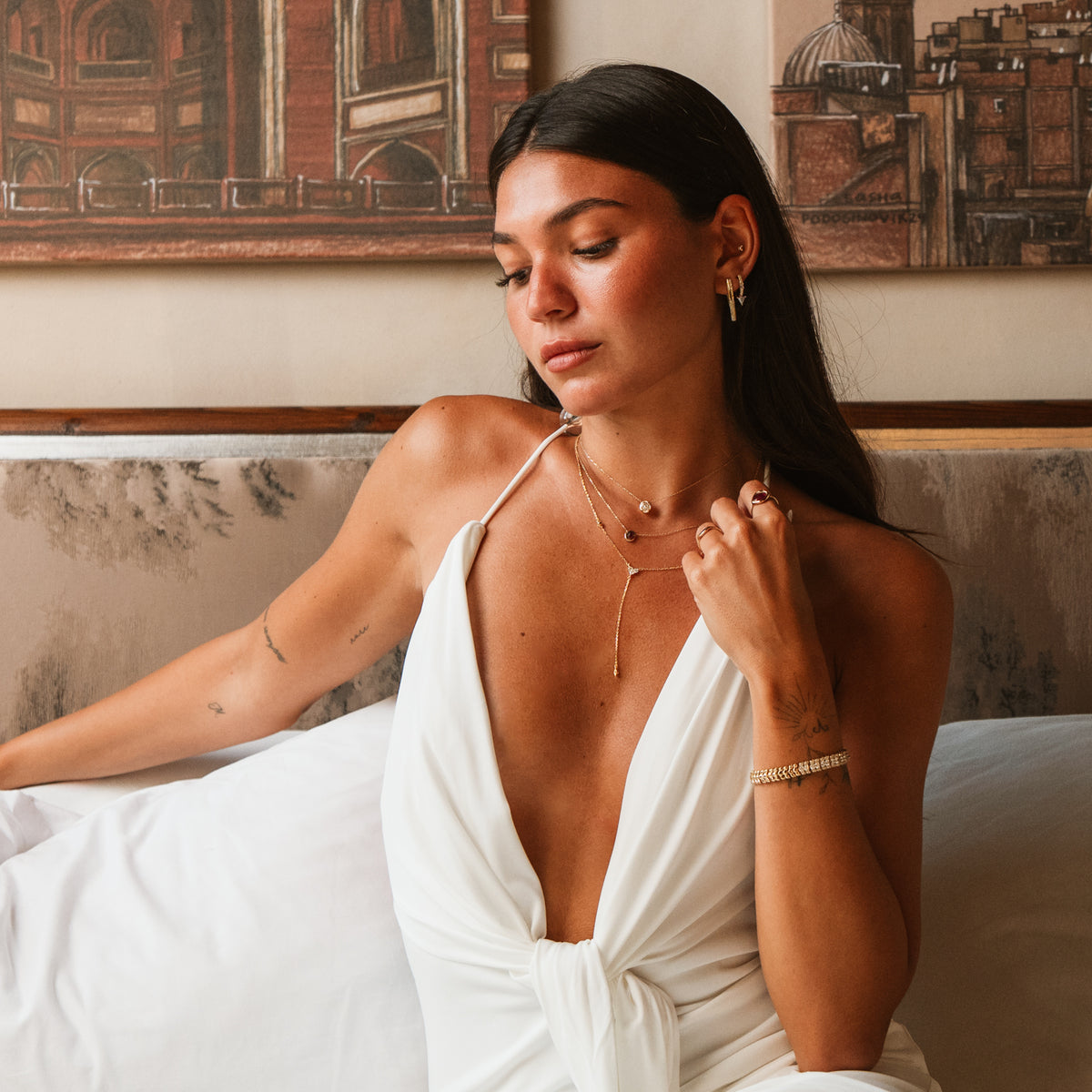 Model seated in a white dress wearing the Trillion Arrow gold stud earrings with layered gold necklaces