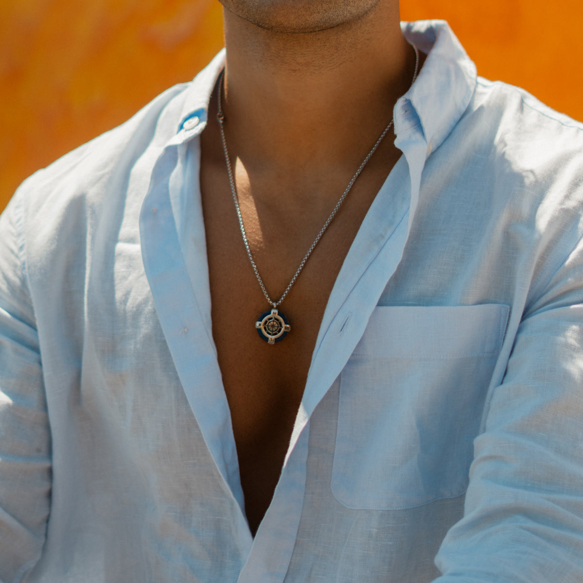 Mens compass necklace worn with an open linen shirt against a sunlit orange wall