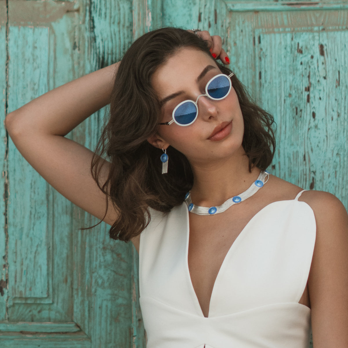 Model with her hand in her hair against a turquoise door, wearing the Cobalt Bloom enamel tassel earring and the matching woven silver choker