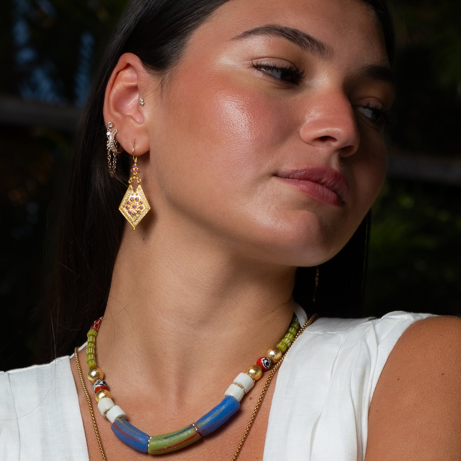 Model wearing Vintage Style Gold & Ruby Design Dangle Earrings by Ebru Jewelry, paired with a colorful beaded statement necklace and a white linen top in a tropical setting.