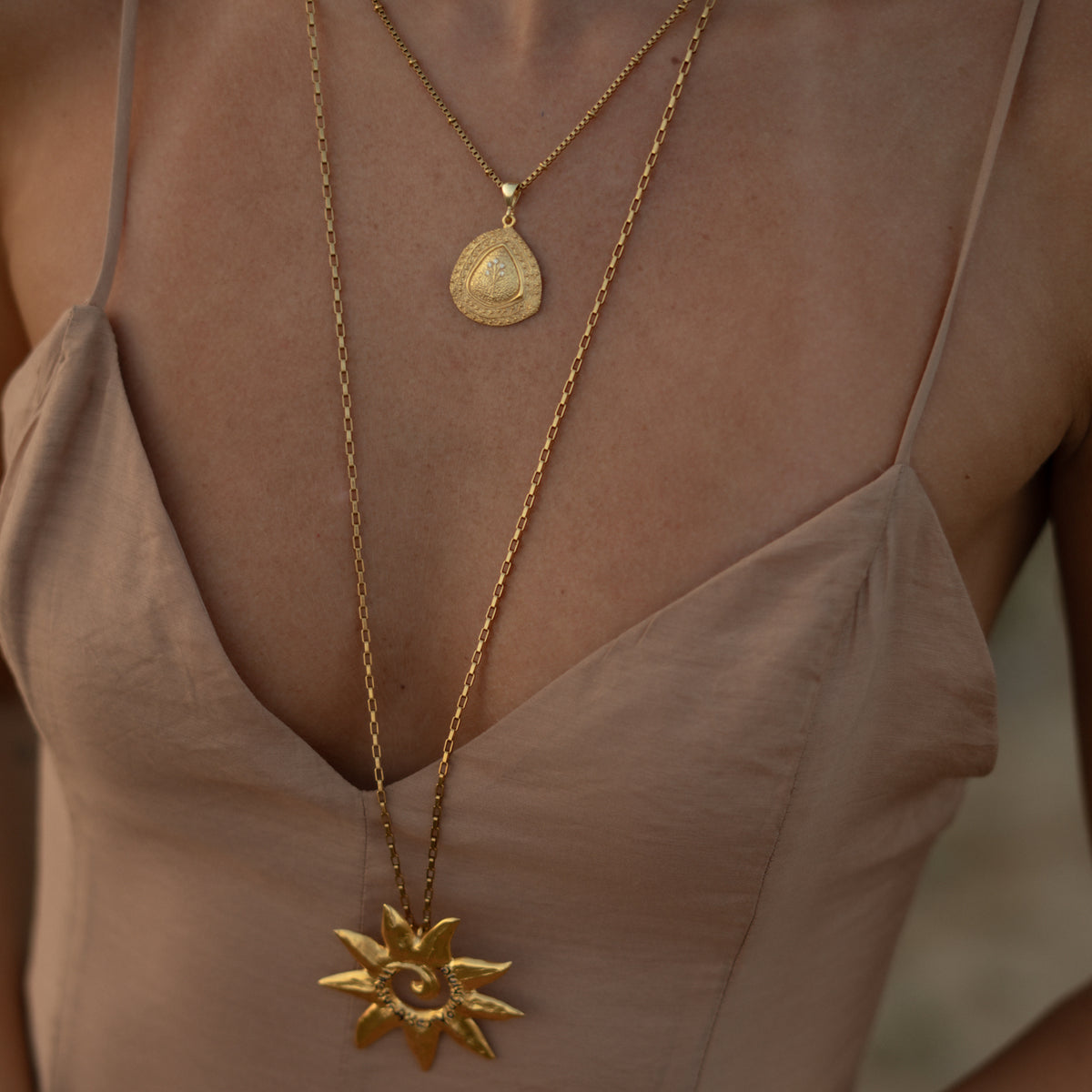 The Tree of Life pendant worn at the collarbone in golden hour light, layered above a long gold sun pendant necklace
