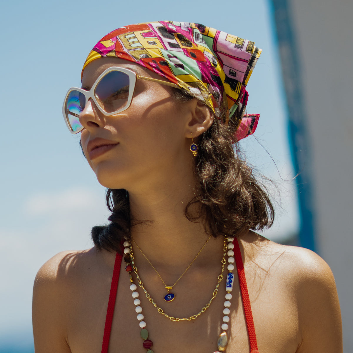 Model in a headscarf and sunglasses wearing the Watch Over Me blue evil eye necklace layered with gold and beaded necklaces