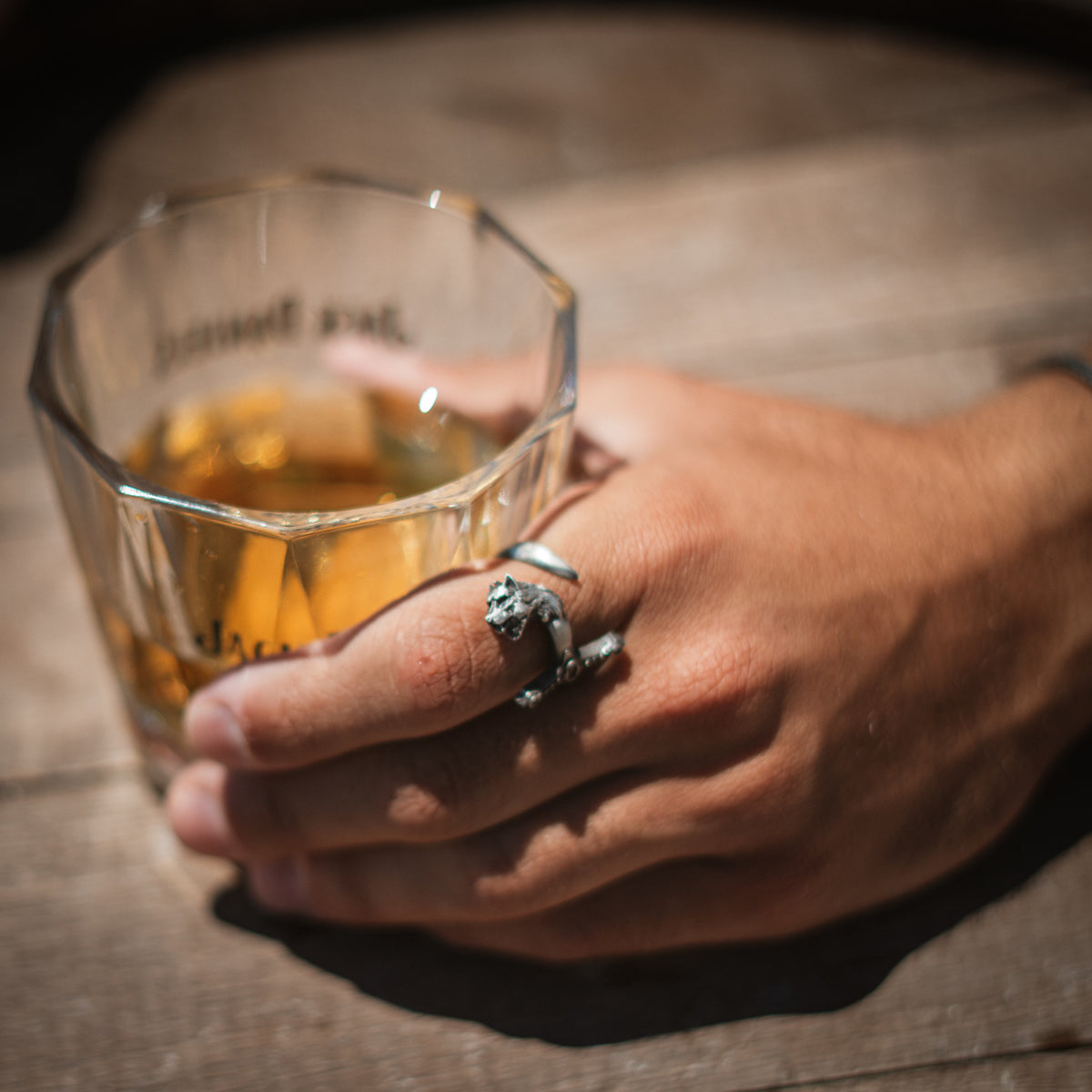 Man's hand resting on a glass at a wooden table, wearing the oxidized sterling silver wolf sword men's ring