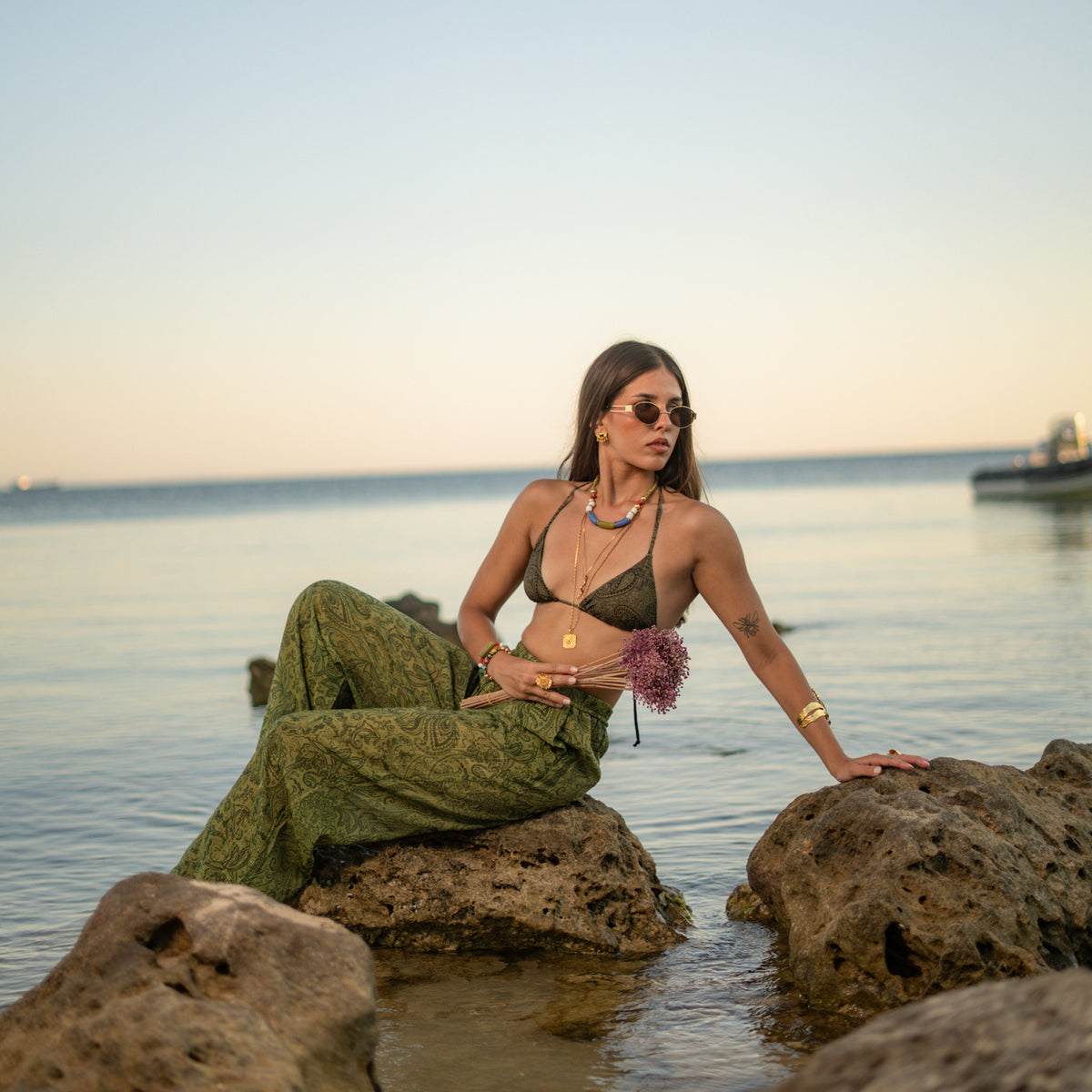 Woman on the rocks at sunset wearing the long gold engraved sun pendant necklace over layered chains