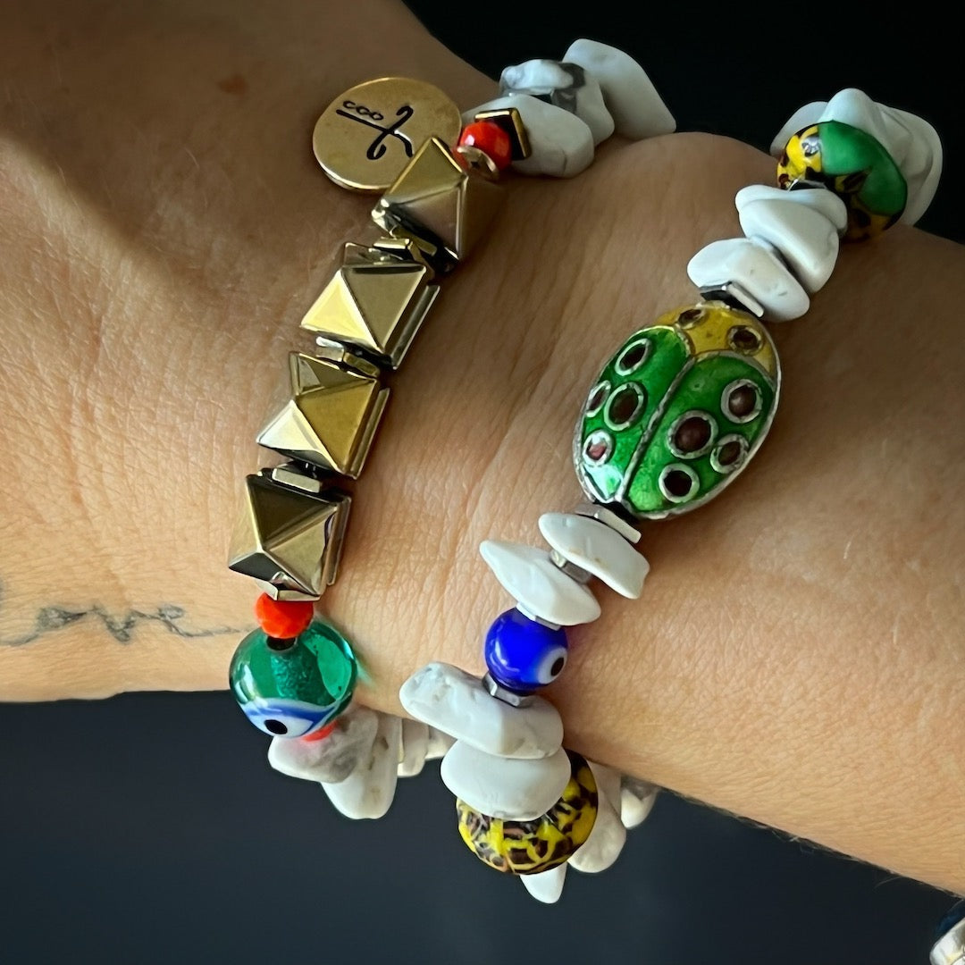 A close-up view of a hand model wearing the Cycle of Life Evil Eye Bracelet. The bracelet&#39;s combination of howlite beads, African beads, hematite stud beads, the evil eye bead, and the bronze heal mantra charm creates a captivating and spiritual piece of jewelry.