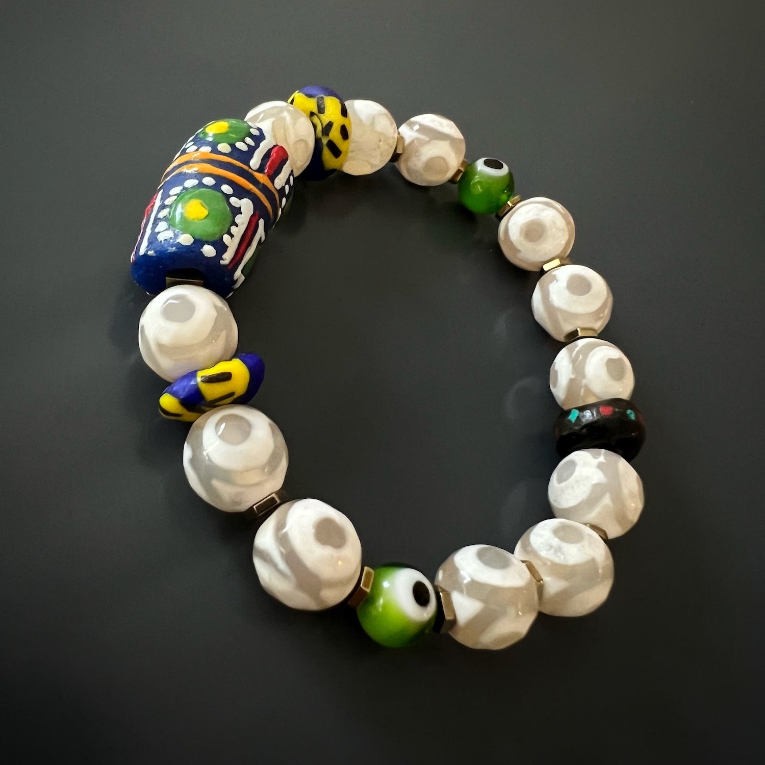 Stay protected and stylish with the Eye of Colors Bracelet, adorned with white agate stone beads, hand-painted African beads, and eye-catching green evil eye beads.