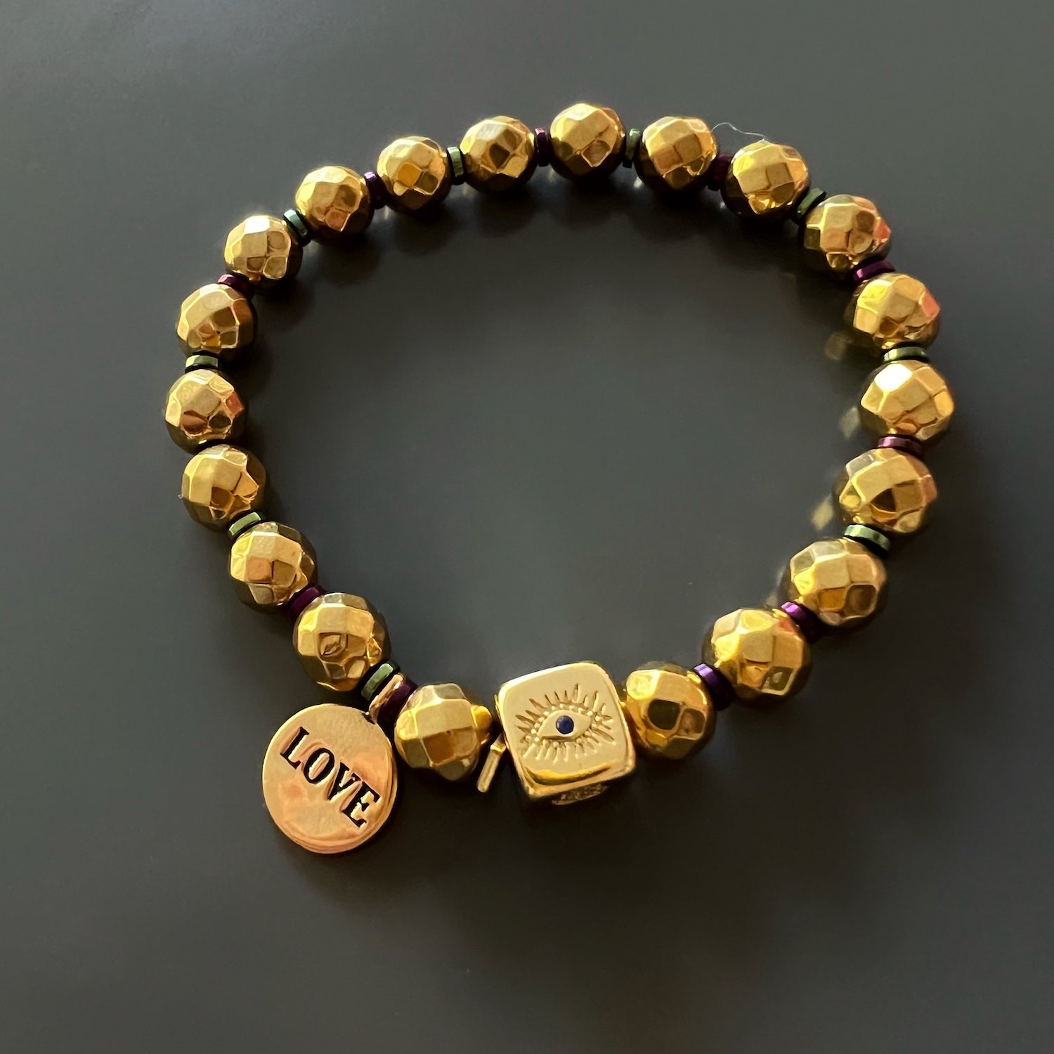 Feel the grounding and protective properties of the Love Symbol Bracelet, featuring a gold Evil Eye and Hamsa accent bead and multicolor hematite spacers.