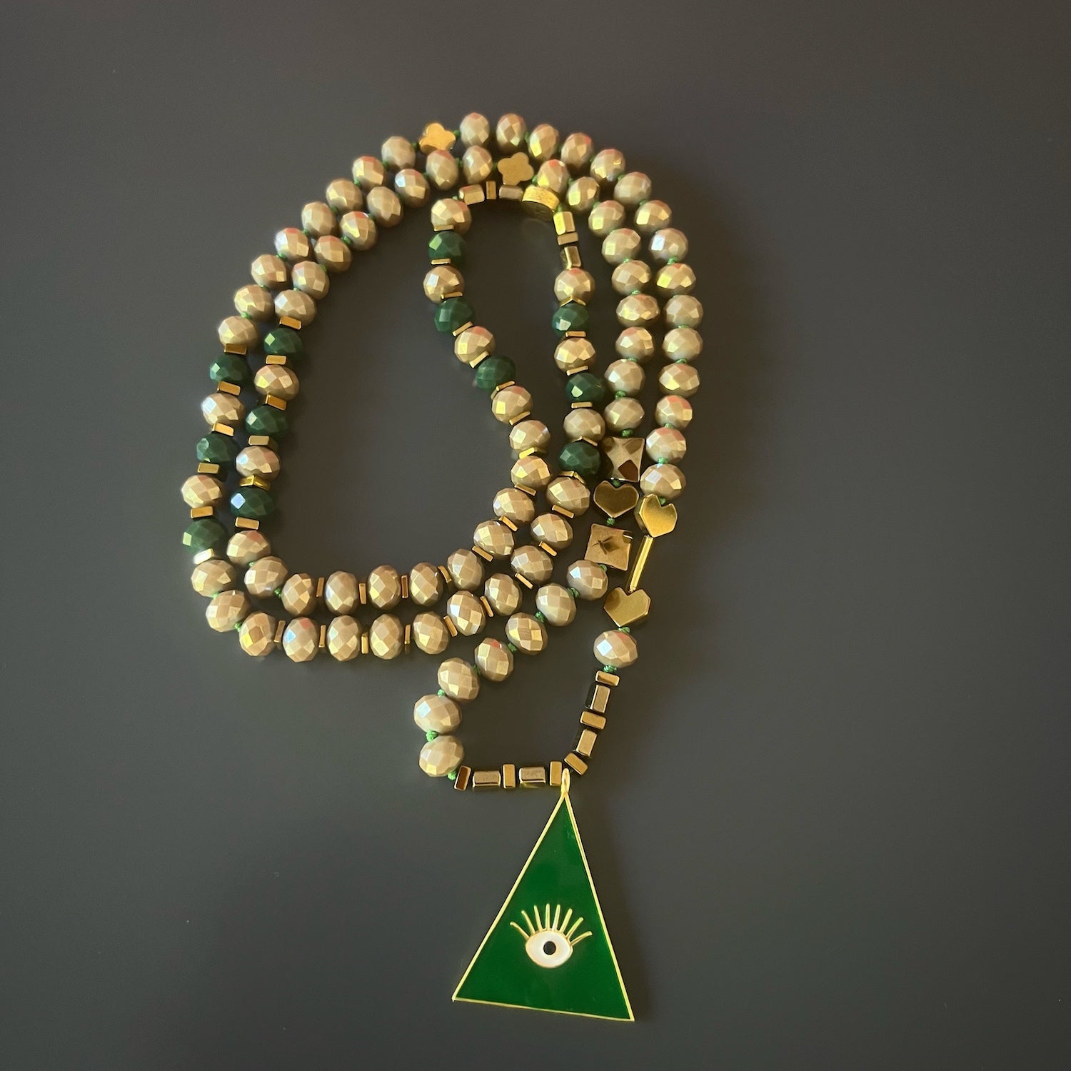 The Green Evil Eye Unique Necklace, elegantly draped around the neck, showcases the harmony of green and beige crystal beads, gold hematite stone heart shape beads, and Alhambra flower beads. 