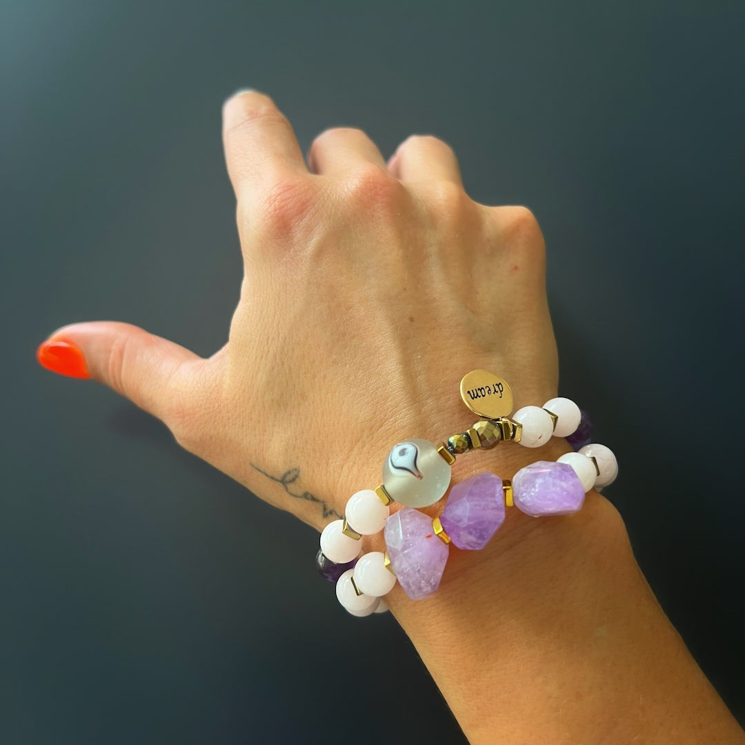 See the Peaceful Mind Bracelet Set adorning the hand model&#39;s wrist, highlighting the soothing properties of rose quartz and amethyst beads, a glass evil eye bead, and a bronze Dream mantra charm.