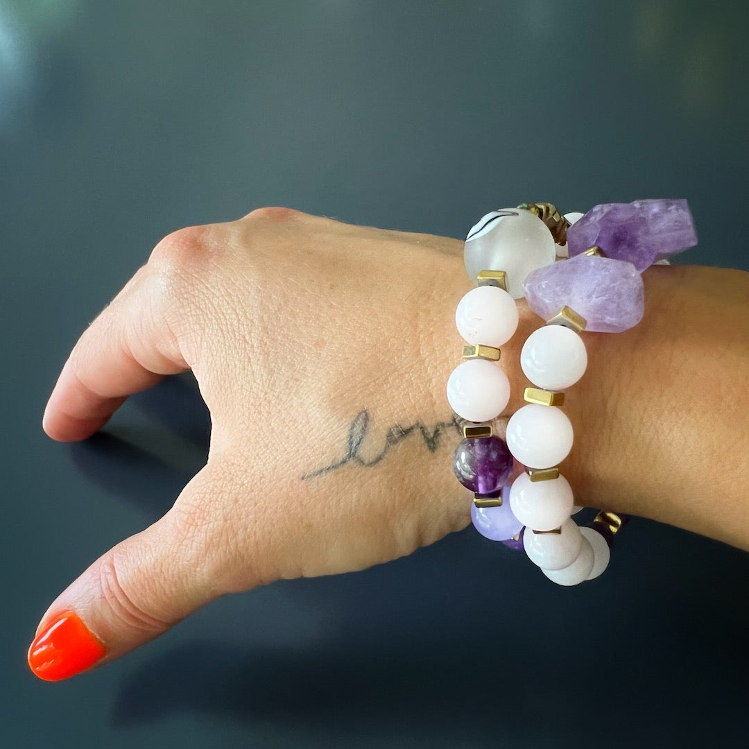 The hand model effortlessly showcases the serenity and balance of the Peaceful Mind Bracelet Set, featuring rose quartz and amethyst beads, a glass evil eye bead, and a bronze Dream mantra charm.