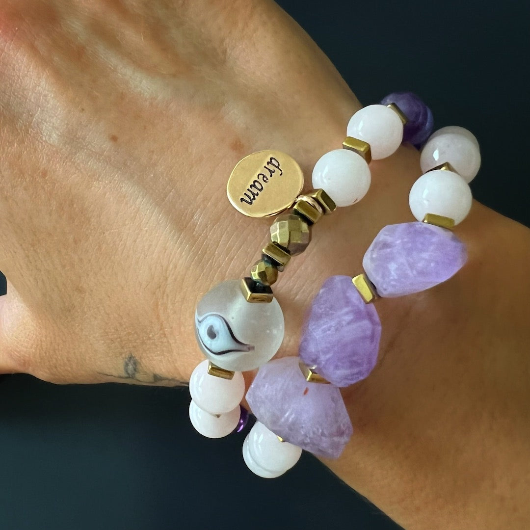 Experience the tranquil energy as the hand model wears the Peaceful Mind Bracelet Set, adorned with rose quartz and amethyst beads, a glass evil eye bead, and a bronze Dream mantra charm.