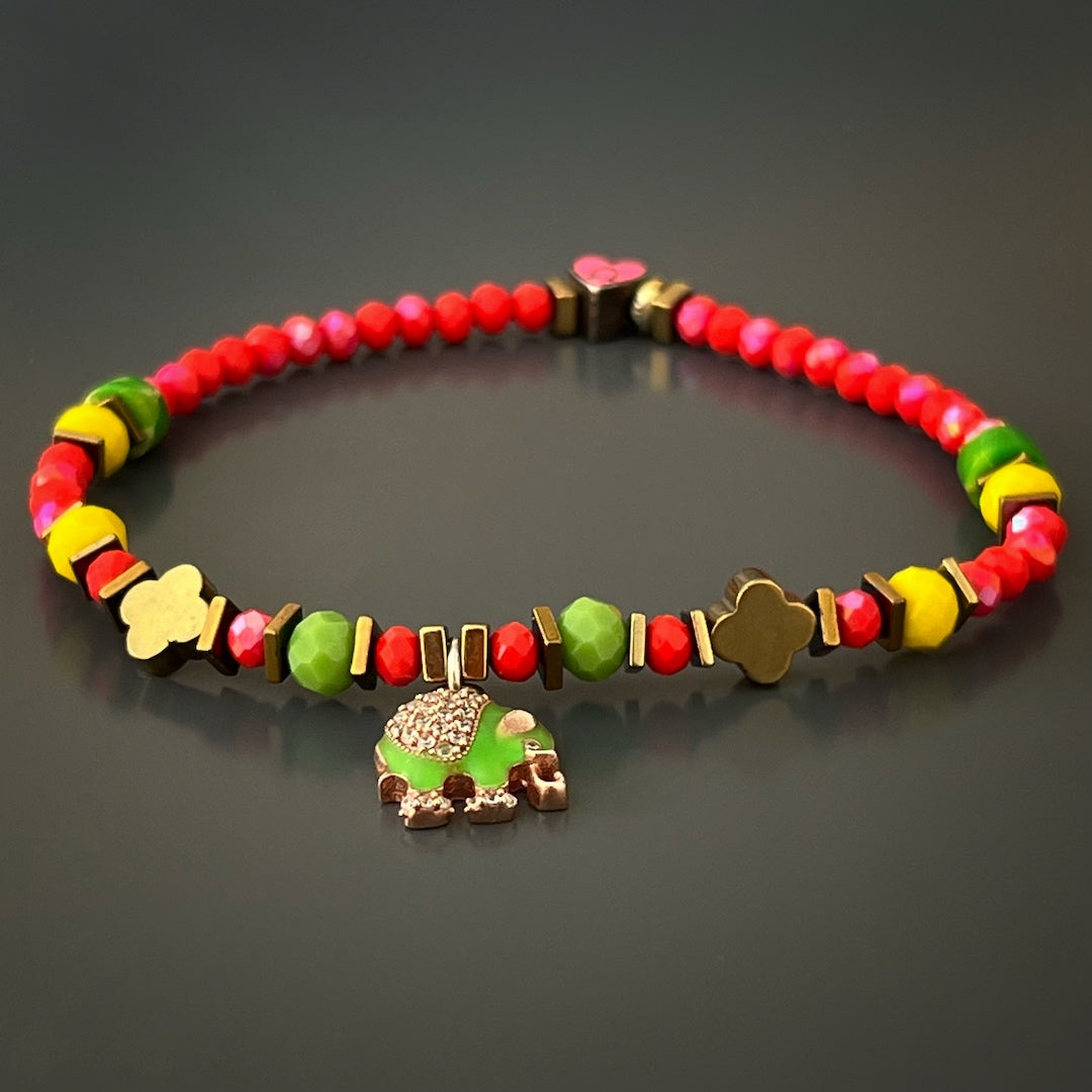 Elevate your style and spirituality with the Powerful Symbol Anklet, showcasing red crystal beads, green jasper beads, a gold color hematite floral bead, and meaningful silver charms symbolizing protection, luck, and strength.