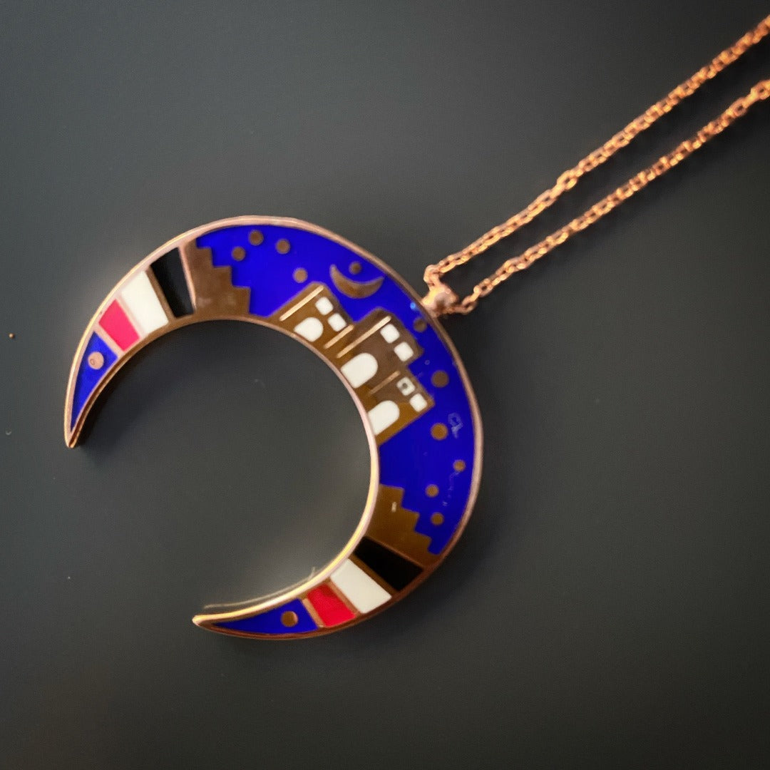 Admire the elegance of the NewYork Necklace, featuring a crescent moon-shaped pendant depicting the colorful New York City skyline in blue, gold, white, and red enamel.