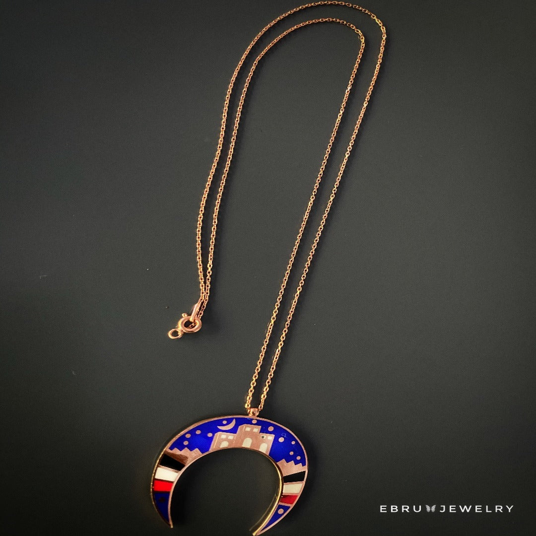 Discover the charm of the NewYork Necklace, showcasing a handmade pendant in the shape of a crescent moon, adorned with a vibrant depiction of the New York City skyline in blue, gold, white, and red enamel.