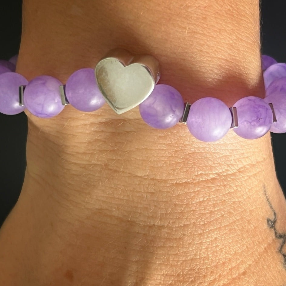 The hand model showcases the beauty and elegance of the Violet Love Bracelet, featuring purple jade beads, sterling silver heart charms, and a sparkling Swarovski crystal accent.